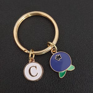 Può includere: Portachiavi color oro con un ciondolo bianco con la lettera "C" e un ciondolo a forma di mirtillo blu con foglie verdi. Il portachiavi è un anello rotondo con due ciondoli pendenti.