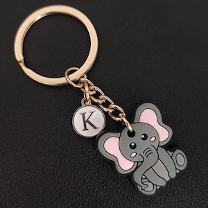 May include: A gold-colored keychain with a gray elephant charm and a white circular charm with the letter "K". The elephant has pink ears and a black outline. The keychain is on a black surface.
