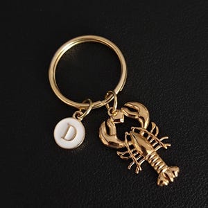 May include: A gold-colored keychain featuring a lobster charm and a white circular charm with the letter "D". The keychain has a large gold ring for attaching keys.