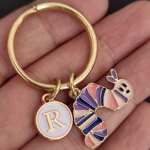 Puede incluir: Llavero dorado con un anillo redondo y dos colgantes. Un colgante es un c&iacute;rculo blanco con la letra "R" dorada. El otro es una oruga colorida con segmentos rosas, morados y azules. La oruga tiene ojos negros y una cara sonriente.