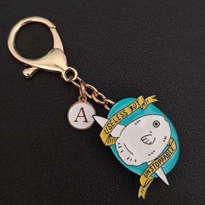 May include: Gold-coloured keychain featuring a cartoon sunfish design. The sunfish is white with a teal background and a yellow banner reading "USELESS BUT UNSTOPPABLE." Includes a charm with the letter "A."