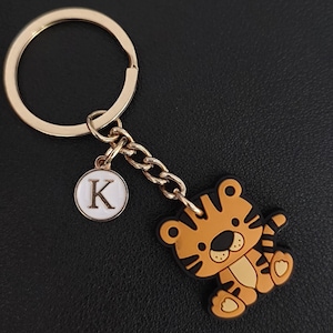 May include: A gold-colored keychain with a cartoon tiger charm and a circular charm with the letter "K". The tiger is orange and black, with a friendly expression. The keychain is on a black surface.
