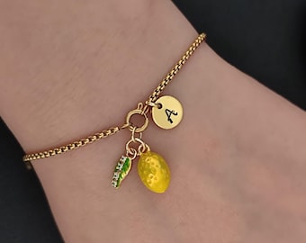 May include: A gold-toned box chain bracelet featuring a circular clasp with three dangling charms: a small round disc engraved with the letter A, a bright yellow lemon charm with a textured surface, and a green leaf charm accented with small clear stones.