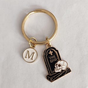 May include: Gold-tone keychain with a white 'M' charm and a black and white tombstone charm. The tombstone reads 'THE END' and features a skull. A unique accessory for keys or bags.