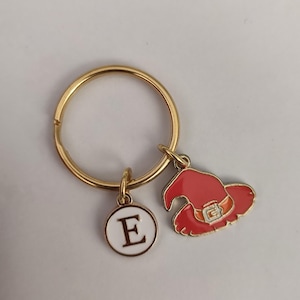 Może przedstawiać: Złoty brelok do kluczy z czerwonym wisiorkiem w kształcie kapelusza czarownicy i białym wisiorkiem w kształcie koła z literą E w kolorze czarnym.