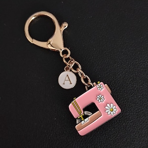 May include: A pink sewing machine charm with daisy accents and a gold-tone clasp. The charm includes a small circular tag with the letter "A". The charm is attached to a gold-tone chain.