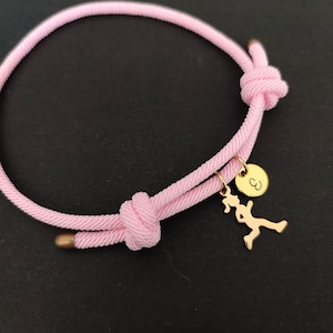 May include: Pink braided rope bracelet with a gold charm of a running figure and a small round charm with the letter "B" engraved on it.
