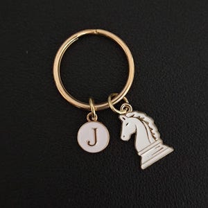 Può includere: Portachiavi color oro con un ciondolo a forma di cavallo degli scacchi in smalto bianco e un ciondolo rotondo con la lettera "J". Il portachiavi è su una superficie nera. Un accessorio elegante per gli appassionati di scacchi.