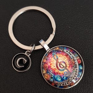 Könnte beinhalten: Silberfarbener Schlüsselanhänger mit einem runden Anhänger, der ein farbenfrohes Musikdesign zeigt. Der Anhänger zeigt einen Violinschlüssel, Klaviertasten und wirbelnde Muster in Rot, Blau und Gelb. Ein kleinerer Charm mit schwarzem Hintergrund und weißem "C" ist ebenfalls angebracht.