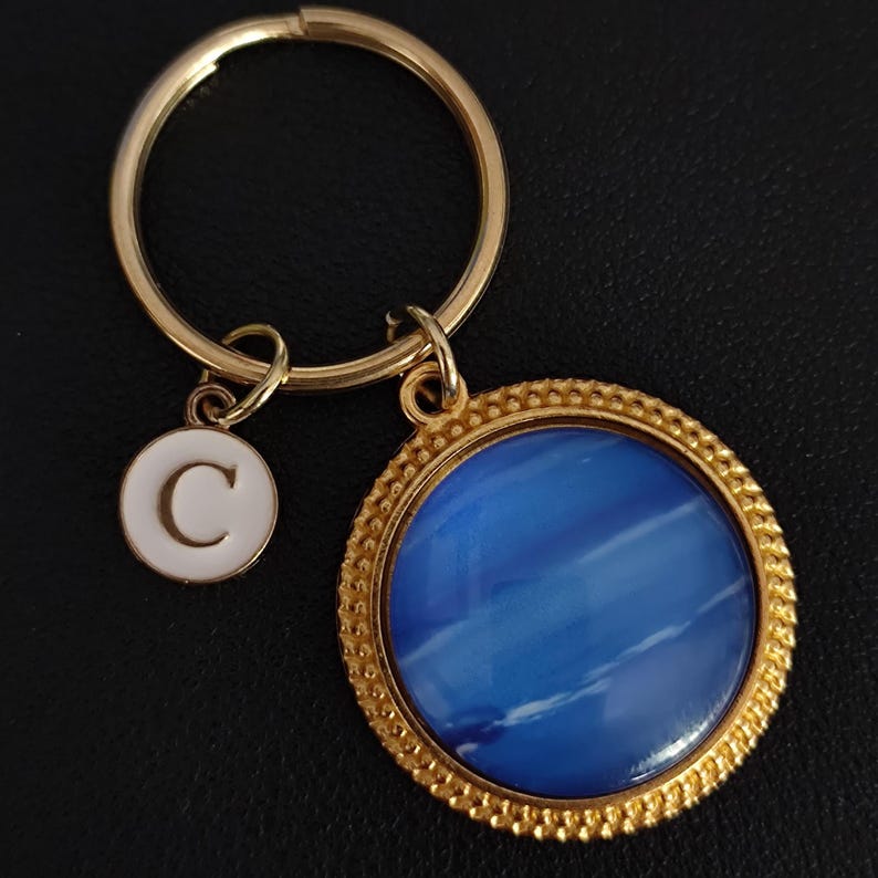 Op de afbeelding: Goudkleurige sleutelhanger met een ronde blauwe hanger en een kleinere witte bedel met de letter "C". De blauwe hanger heeft een getextureerde uitstraling en de sleutelhanger is aan beide bedels bevestigd.