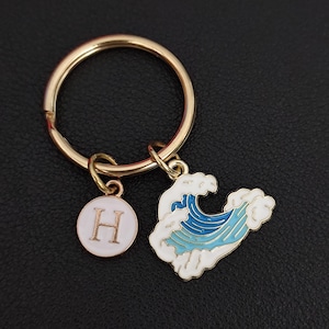 May include: Gold-tone keychain with two charms. One charm is a white circle with the letter "H" in the center. The other charm is a blue and white wave design. The keychain is a stylish accessory.