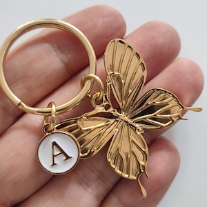 Può includere: Portachiavi a forma di farfalla dorata con un ciondolo rotondo bianco con la lettera A in oro.
