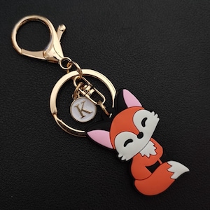 Könnte beinhalten: Ein goldfarbener Schlüsselanhänger mit einem Cartoon-Fuchs-Anhänger. Der Fuchs ist orange, weiß, schwarz und rosa. Ein kleiner, runder Anhänger mit dem Buchstaben "K" hängt am Schlüsselanhänger.