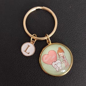 Op de afbeelding: Goudkleurige sleutelhanger met een ronde bedel met een tandheelkundig ontwerp en een kleinere bedel met de letter "L". De tandheelkundige bedel heeft een cartoon tandarts, een tand en een hart met de woorden "Dental Care!"