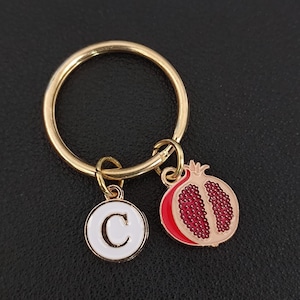 May include: Gold-coloured keyring with a white enamel charm featuring the letter "C" and a red and tan enamel pomegranate charm. A decorative accessory.
