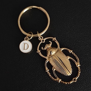 May include: Gold-colored keychain featuring a detailed beetle pendant and a circular charm with the letter "D" in white. The keychain includes a large ring for keys and a smaller ring to attach the charm and beetle.