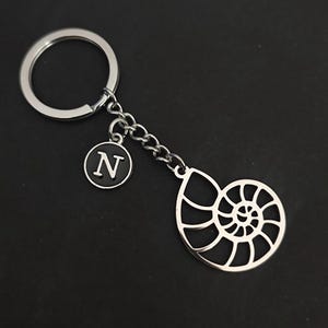 Op de afbeelding: Zilverkleurige sleutelhanger met een nautilusschelpontwerp. De sleutelhanger bevat een ketting, een ronde bedel met de letter "N" in een zwarte cirkel en een sleutelring. De achtergrond is zwart.