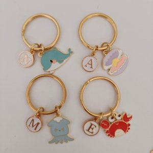 Op de afbeelding: Vier goudkleurige sleutelhangers met zeethema bedels. Bedels zijn onder meer een blauwe walvis, een schelp, een krab en een blauwe octopus. Elke sleutelhanger heeft een letterbedel: N, A, M en E. Op een witte achtergrond.