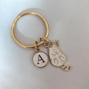 Könnte beinhalten: Goldener Schlüsselanhänger mit einem weißen Katzenanhänger und einem goldenen Kreisanhänger mit dem Buchstaben A.