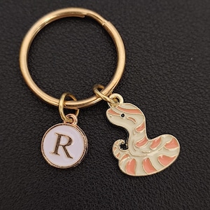 May include: Gold-coloured keyring with two charms: a white and pink snake and a round charm with the letter "R" on a white background. The keyring is on a black surface.