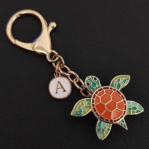May include: Gold-toned keychain featuring a turtle charm with a red and orange shell, green and teal flippers, and a small white disc with the letter "A". The keychain has a lobster clasp and a chain.