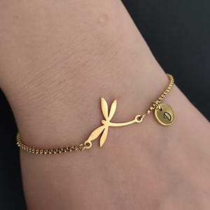 Könnte beinhalten: Goldfarbenes Armband mit einem Libellen-Anhänger und einem kleinen runden Anhänger mit dem Buchstaben "D". Das Armband hat ein filigranes Ketten-Design. Der Libellen-Anhänger ist eine stilisierte Silhouette.