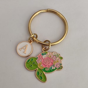 May include: Gold-coloured keyring featuring a floral turtle charm and a white circular charm with the letter "A". The turtle charm is green with pink and white flowers. A decorative accessory.