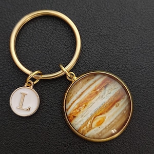 Könnte beinhalten: Ein goldfarbener Schlüsselanhänger mit zwei Anhängern. Ein Anhänger ist ein runder Anhänger mit einem detaillierten Bild des Jupiter. Der andere ist ein kleiner, weißer, runder Anhänger mit dem Buchstaben "L". Der Schlüsselanhängerring ist ebenfalls goldfarben.