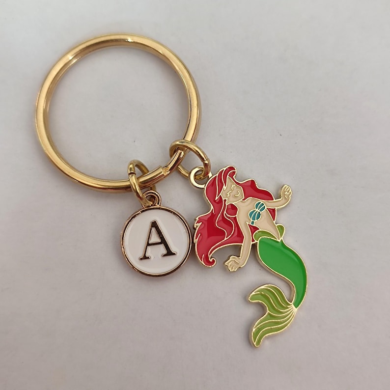 May include: Gold key ring with a white circle charm featuring the letter A and a red and green mermaid charm.