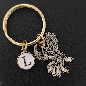 May include: Gold-coloured keyring with a round charm bearing the letter "L" and a detailed phoenix pendant. The phoenix is black and white with gold accents. The keyring is on a black surface.