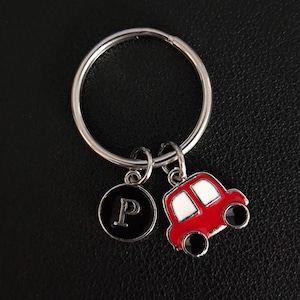 Può includere: Un portachiavi argentato con un ciondolo a forma di auto rossa e un ciondolo circolare nero con la lettera "P" in bianco. Il ciondolo dell'auto ha finestrini bianchi e ruote nere. Il portachiavi è su una superficie nera.