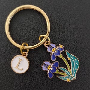 May include: Gold-coloured keyring with a white circular charm bearing the letter "L" and a floral charm depicting blue irises. The iris charm has green leaves and yellow star details. The keyring is on a black surface.