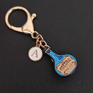 Könnte beinhalten: Ein goldfarbener Schlüsselanhänger mit Karabinerverschluss, einem weißen runden Anhänger mit dem Buchstaben "A" und einem blauen Flaschenanhänger. Der Flaschenanhänger trägt die Aufschrift "Happily Ever After" und "True Love".