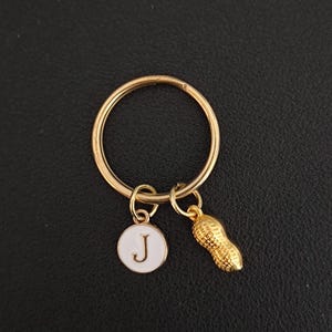 Könnte beinhalten: Goldfarbener Schlüsselanhänger mit rundem Schlüsselring. Der Anhänger hat zwei Anhänger: einen weißen, kreisförmigen Anhänger mit dem Buchstaben "J" und einen goldenen Erdnuss-Anhänger. Der Hintergrund ist dunkel und strukturiert.