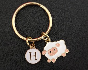 May include: Gold-colored keychain with a round ring and two charms. One charm is a white circle with the letter "H" in the center. The other charm is a white sheep with pink accents.