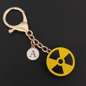 Może przedstawiać: Złoty brelok do kluczy z zawieszką w kształcie symbolu promieniowania i białą okrągłą zawieszką z literą "A". Symbol promieniowania jest żółty i czarny. Brelok ma zapięcie na karabińczyk.