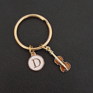 Puede incluir: Un llavero dorado con un anillo redondo y dos colgantes. Un colgante es un círculo blanco con la letra "D" en el centro. El otro colgante es un violín en miniatura marrón y dorado.