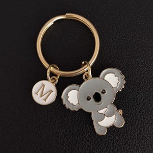 Peut inclure: Un porte-clés en métal doré avec un charm en forme de koala gris et blanc et un charm rond avec la lettre "M". Le koala a des yeux noirs et est en position debout. L'anneau du porte-clés est également doré.