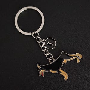 May include: A silver keyring with a black and tan dog pendant. The dog is depicted in a playful stance with its tongue visible. A small, circular charm with the letter "I" is also included. The keyring has a silver ring and chain.