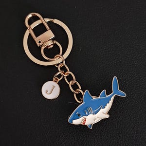 May include: Gold-coloured keychain with a blue and white shark charm with red accents. Includes a small white circular charm with the letter "J". The keychain has a lobster clasp and a split ring.