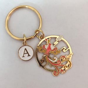 May include: Gold-tone keyring with a round charm featuring a red and white phoenix with a gold and yellow tail. The charm is attached to a gold-tone key ring with a smaller round charm that says "A" in white letters on a white background.