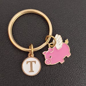 May include: Gold key chain with a pink pig charm with white wings and a white circle charm with the letter T in gold.