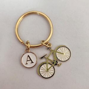 Peut inclure: Porte-clés en or avec un charme de vélo vert et un charme de cercle blanc avec la lettre A en noir.