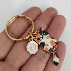 May include: Gold keychain with a calico cat charm and a white circle charm with the letter N in gold. The cat charm has a black tail with white and orange stripes.