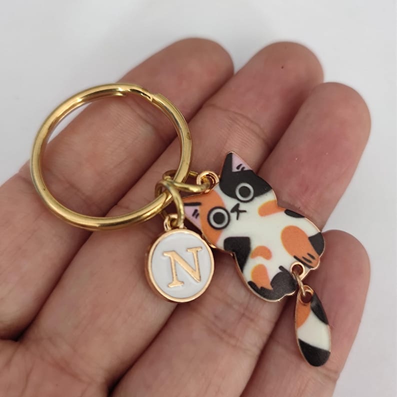 May include: Gold keychain with a calico cat charm and a white circle charm with the letter N in gold. The cat charm has a black tail with white and orange stripes.