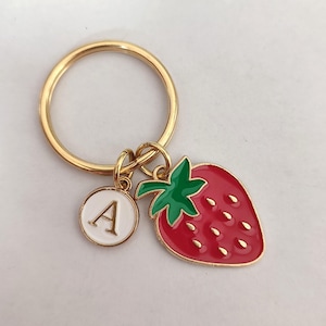 Op de afbeelding: Een goudkleurige sleutelhanger met een rode aardbei-bedel en een witte cirkelvormige bedel met de letter "A". De aardbei heeft groene bladeren en goudkleurige zaadjes. De sleutelhanger staat op een witte achtergrond.
