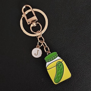May include: Gold-tone keychain with a charm featuring a jar of pickles with a smiling face. The keychain also includes a small white charm with the letter "J".