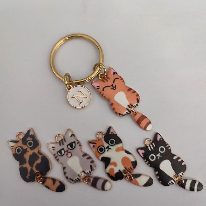 May include: Five colorful cat keychains with gold rings. Each keychain features a different cat design with a black and white striped tail. The keychains are on a white background.