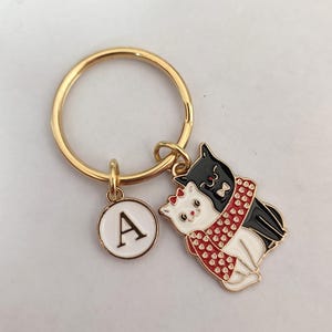 May include: Gold-coloured metal keyring with a charm depicting two cats, one black and one white, embracing and wearing a red scarf. A white circular charm with the letter "A" is also attached.