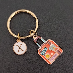 Könnte beinhalten: Goldfarbener Schlüsselanhänger mit rundem Verschluss, einem kreisförmigen Anhänger mit dem Buchstaben "X" und einem roten Kofferanhänger. Der Kofferanhänger trägt die Wörter "TRAVEL" und "HELLO", eine Palme und ein Smiley.